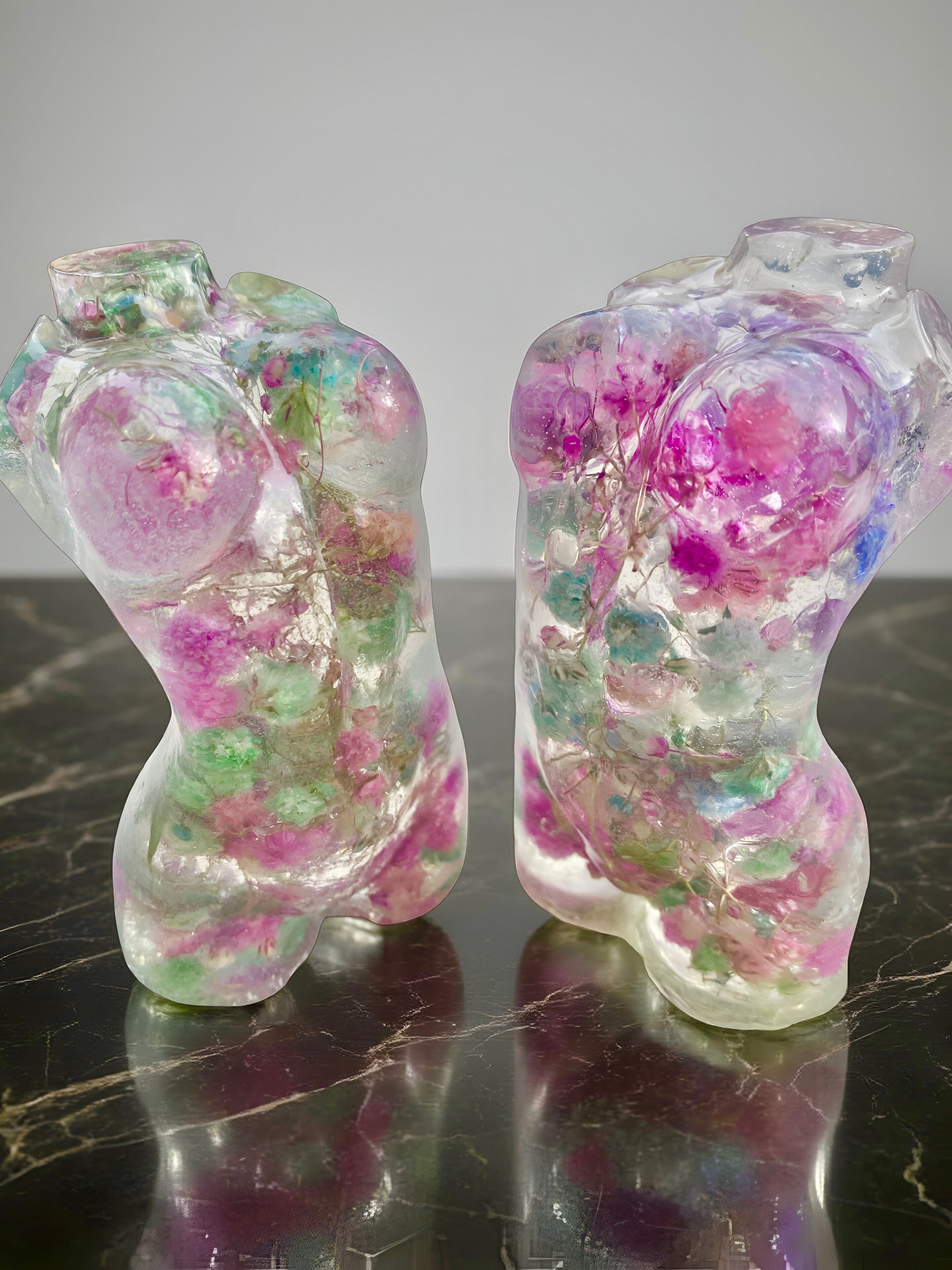 Two translucent resin women's torsos with colorful embedded materials on a dark surface.